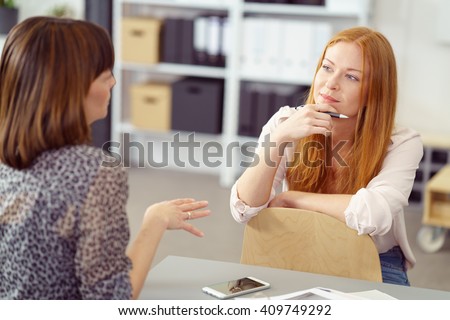 Two businesswoman having an informal meeting with one sitting relaxing on a reversed chair listening to her colleague with a pensive expression Royalty-Free Stock Photo #409749292