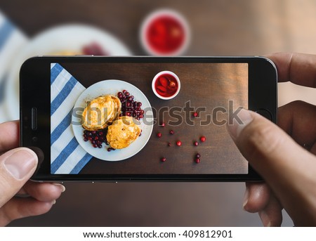Taking picture of pancakes with mobile phone. Phone in male hands.On the plate there is pancakes with berries and honey. Plate is on a black aged wooden table. Vintage style.