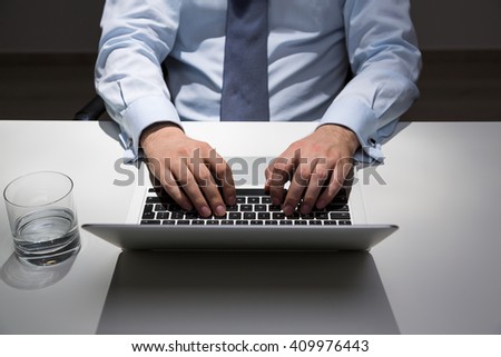 Businessman sitting at office desk with glass of water and typing on laptop keyboard. Spot lighting