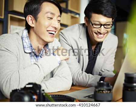 two asian photographers working together selecting images using laptop computer, happy and laughing.