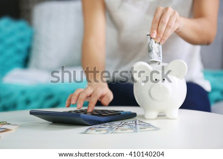Female hand putting money into piggy bank and counting on calculator closeup Royalty-Free Stock Photo #410140204