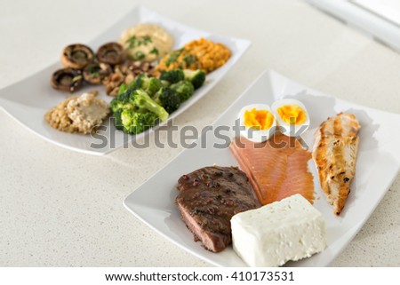 animal versus plant proteins: one plate with beef, eggs, salmon, cheese and chicken grill and another with nuts, mushrooms, broccoli, lentil, hummus and quinoa Royalty-Free Stock Photo #410173531