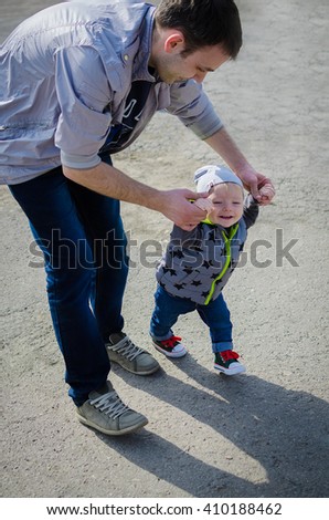 Father guiding his son with the first steps outside. Baby learning to walk, holding father's hands. Toddler steps with the help of his father in spring park. Stylish boy in jeans and sneakers.