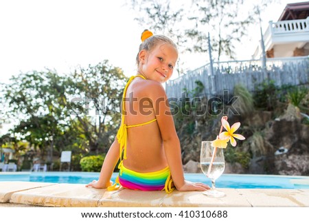 Cute little blonde girl swimming in big pool. Big glass with water, straw and frangipani stay on the pool edge. Little lady sit and look to the camera. Sunbathing and leisure on sunny summer day.
