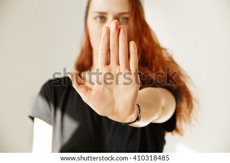 Close up view of young woman making stop gesture with her hand. Cropped isolated portrait of redhead female in cap and black T-shirt standing against white background.