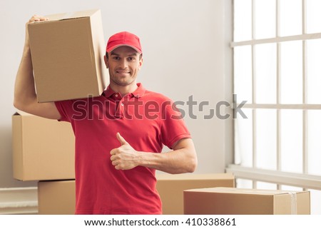 Handsome delivery worker is showing Ok sign, holding a box, looking at camera and smiling while standing among cardboard boxes