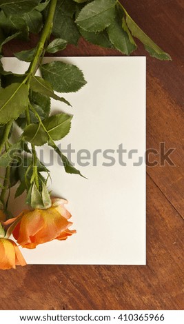 A photo of a blank white card shot from above, with some orange colored roses, on a wooden background texture, with copyspace