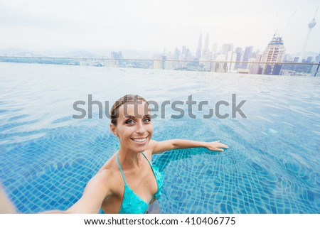 Vacation and technology. Pretty young woman taking selfie while swimming in roof top pool with beautiful city view.