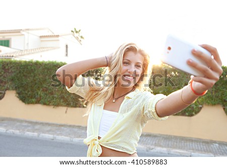 Portrait of a beautiful blond teenager using a smart phone to network, taking selfies pictures of herself in a suburban home exterior at sunset, outdoors. Technology lifestyle, adolescent activities.
