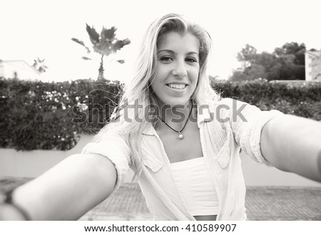 Black and white portrait of a beautiful teenager girl holding a smart phone taking selfies photos and videos in a suburban home exterior, outdoors. Technology lifestyle, adolescent smiling at camera.