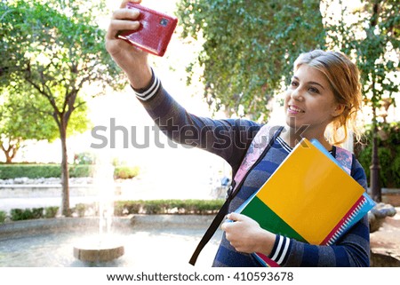 Portrait of beautiful student teenager girl holding up smartphone taking selfies, networking using technology, outdoors park. Adolescent with books taking photos by water fountain, lifestyle exterior.