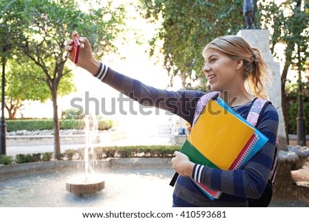 Beautiful student teenager girl holding up a smartphone taking selfies of herself, networking using technology, outdoors park. Adolescent with books taking photos, water fountain lifestyle exterior.