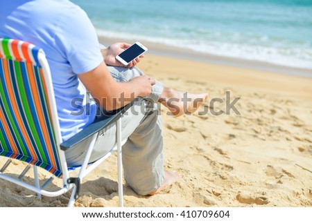 Back side view of man relaxing on the beach outdoors background using smartphone. Barefoot luxury relaxation