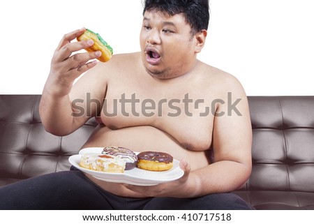 Picture of a young obese man sitting on brown sofa and enjoy donuts, isolated on white background