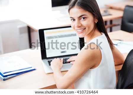 Young pretty businesswoman using laptop computer in office and looking back at camera
