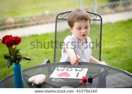 Little 7 year old boy paints greeting card for Mom on Mother's Day with the inscription "I love you mom". Outdoors. Mother's Day