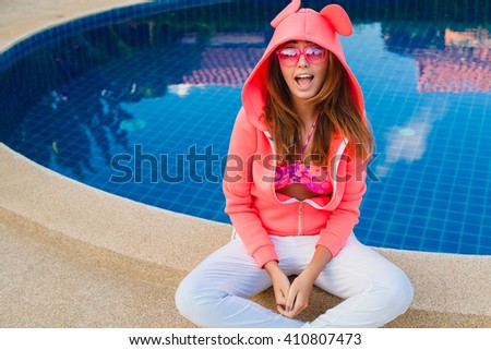 young hipster woman sport style sitting at pool, pink neoprene hoodie, stylish apparel, sunglasses, fun, summer, funny face, expression, laugh, smiling, positive, crazy