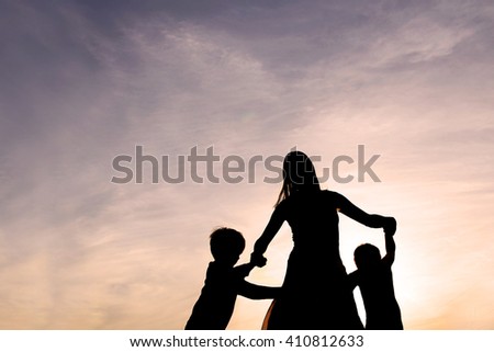 Silhouette of a mother and her two young children holding hands and dancing around outside, isolated against the sunset. Royalty-Free Stock Photo #410812633