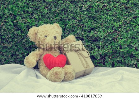 Brown teddy bear sitting on white fabric with red heart yarn and gift box. Tree leaf bushes green fence, Texture background.