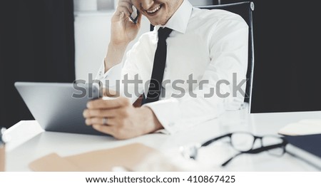 Happy smiling businessman working at his modern office on digital tablet and talking on smartphone