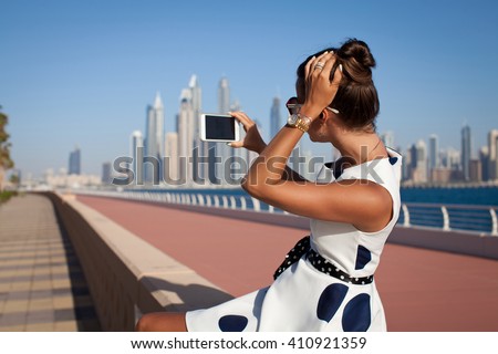 Girl holding smartphone for self-portrait photo with view of Dubai skyscrapers during summer travel vacation.