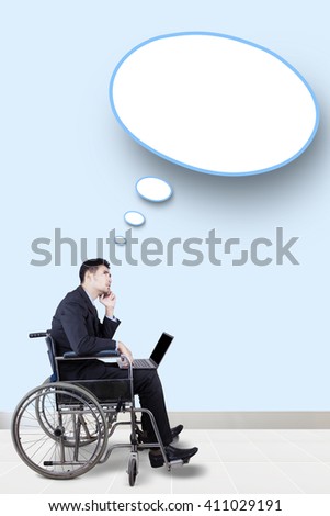 Picture of disabled businessman sitting on the wheelchair with a laptop and looking at empty speech bubble