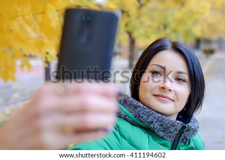Gorgeous young woman in thick winter coat taking picture of herself at park with camera phone under yellow tree leaves in background