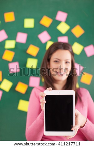 Young woman standing in front of chalkboard with scheme with stickers in different colors, and holding a digital tablet and showing it. She is looking a the camera and smiling 