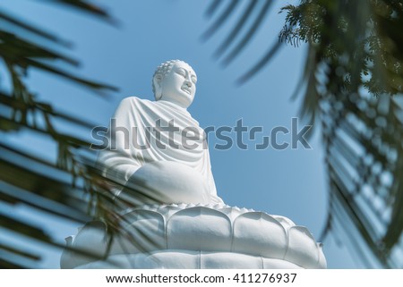 Horizontal picture of white statue of Buddha in Long Son Pagoda in Nha Trang, Vietnam, sitting on lotus by background of blue sky (view through green leaves of palm trees)
