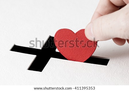 Red heart symbol is put by person's hand into slot of white donation box, slot is shaped like Christian cross. Concept of sincere devotion to faith with whole heart. Close-up shot Royalty-Free Stock Photo #411395353