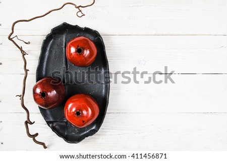 Three ceramic red pomegranates and black plate on white wooden background. Flat lay set of color  house decor. Organic food, pottery, decoration concept. Bright ceramic red pomegranates on black dish
