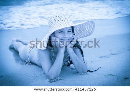 Smiling girl in pink hat on the beach lying down on the sand near water, blue toned picture