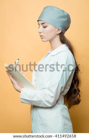 Female doctor writes on paper plate - a folder with medical documents in white uniform in studio on orange background