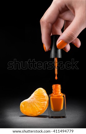 Juicy nail treatment with bright orange colors of nail polish and citrus' segment near it. Black background. Female hand with orange nails is holding the brush. Positive picture.
