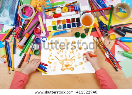 child drawing baby stuff - pram, wear and toys, top view hands with pencil painting picture on paper, artwork workplace