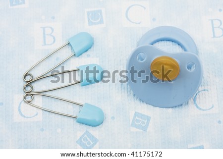 Blue diaper pins and pacifier sitting on a blue alphabet background, diaper pin and rattle