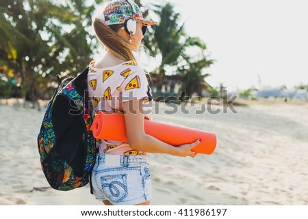 young beautiful woman walking on beach with yoga mat, listening to music on headphones, hipster sport swag style, denim shorts, t-shirt, backpack, cap, sunny, summer weekend, view from back