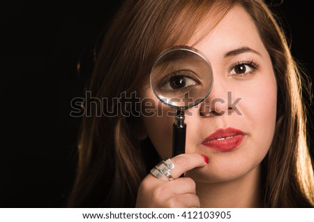 Young woman facing camera holding magnifying glass over right eye creating enhanced effect, black background