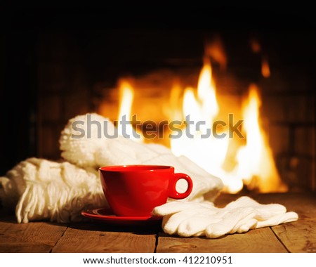 Red cup of coffee or tea, woolen scarf, gloves and cap on wooden table near  fireplace. Winter and Christmas holiday concept. 