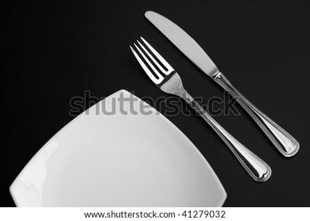 Knife, square white plate and fork on black background
