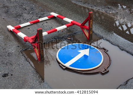 Overturned construction safety barrier and turn right sign in the puddle on the road. The sign is on the manhole cover  inside the cut out part of asphalt surface prepared for paving with new asphalt.
