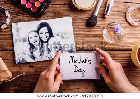 Black-and-white picture of mother hugging her daughters and various make up products. Hands of unrecognizable woman holding Mothers Day card. Studio shot on wooden background.