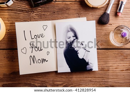Mothers day composition. Black-and-white picture of mother holding her little baby, greeting card with I love you Mom text and various cosmetics. Studio shot on wooden background.