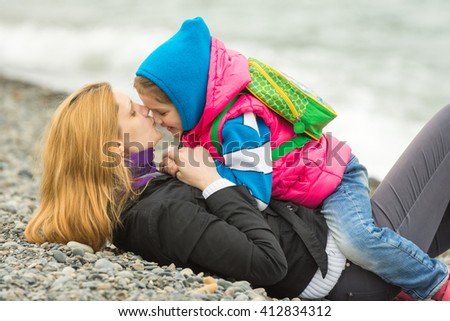 Young mother lying on the pebbly seashore kissing the nose her daughter sitting on her riding