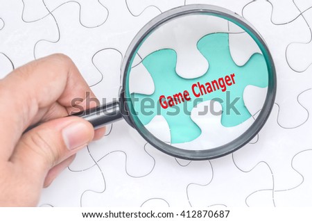 Game Changer word with hand holding magnifying glass over jigsaw puzzle. Selective focus.