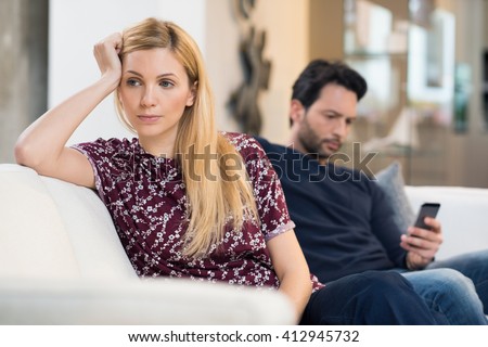 Young woman getting bored while man using phone in the background. Beautiful young woman feeling annoyed as man texting on phone. Young woman after an argument with her boyfriend in their living room. Royalty-Free Stock Photo #412945732