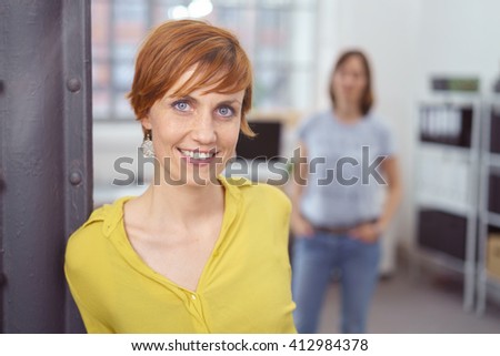 Cute young red haired woman in yellow blouse leaning against post in small office with partner or friend out of focus in background
