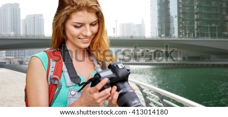 adventure, travel, tourism, hike and people concept - happy young woman with backpack and camera photographing over dubai city waterfront and bridge background