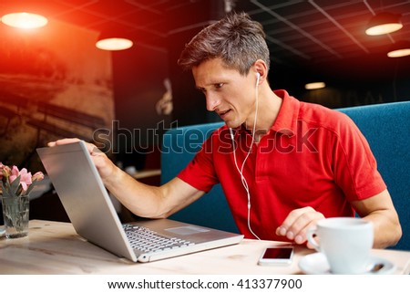 joyful smiling man sitting in a cafe near the open laptop at a table made of wood.  in the background a bright window with bright daylight