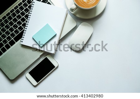 Coffee cup, smart phone, notepad, and laptop on white table. View from above
